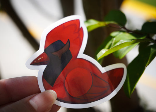 Red bird-shaped sticker held by a hand with blurred green leaves in the background