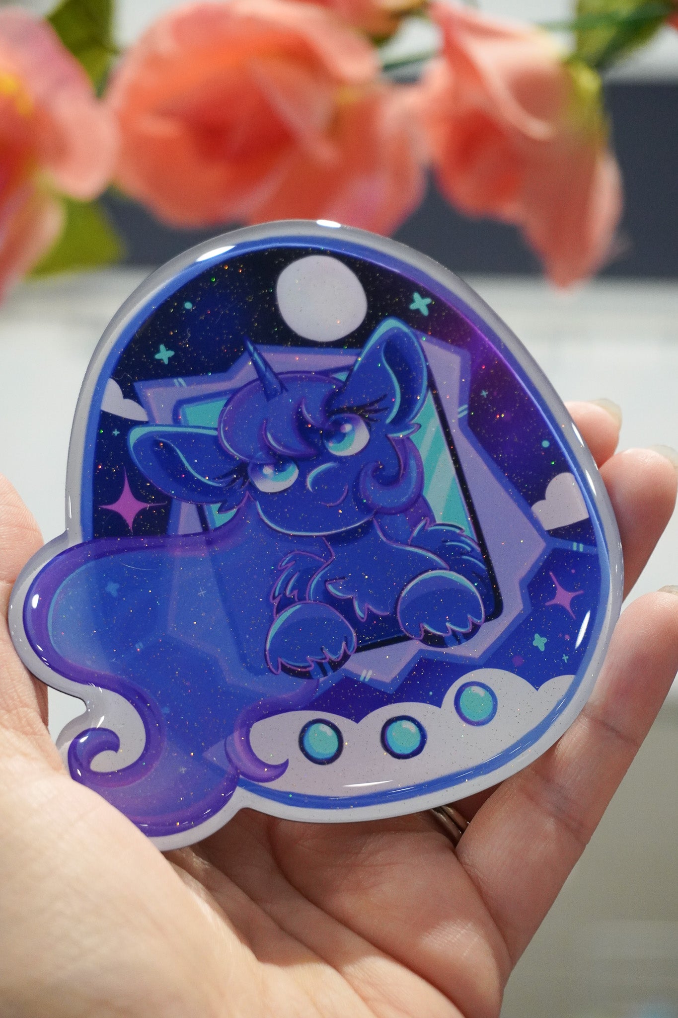 Sticker with a blue pony character design held by a hand, with flowers in the background