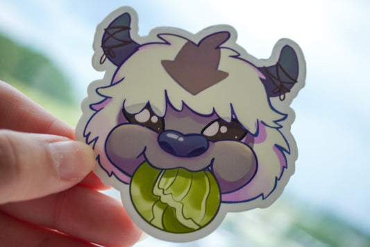 Sticker of a cartoon character with horns and a green ball held by a hand against a blurred outdoor background.